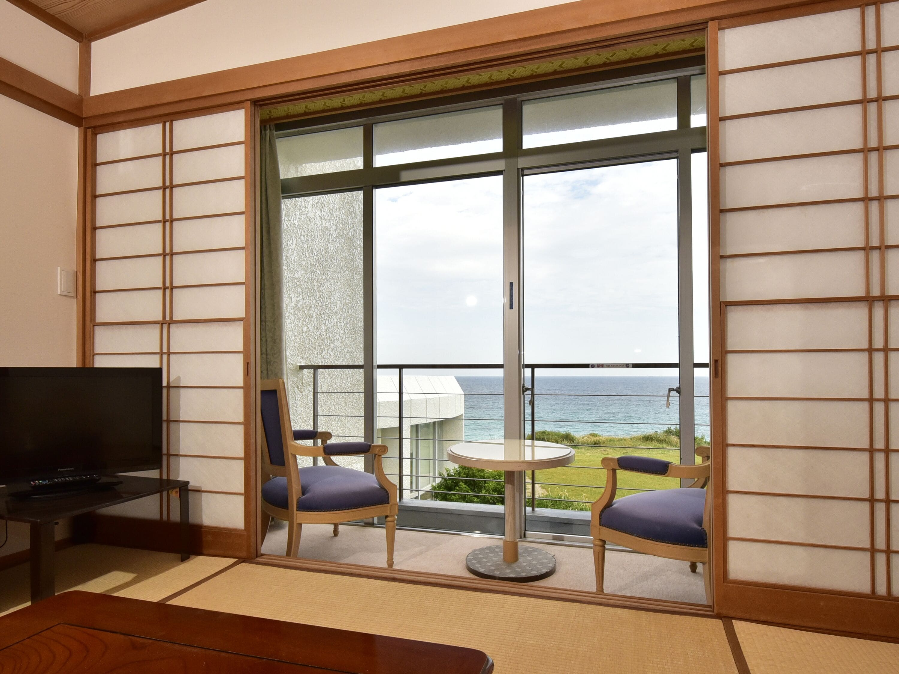 A room with a television, two chairs, a small table, and a view of the sea through large glass doors.