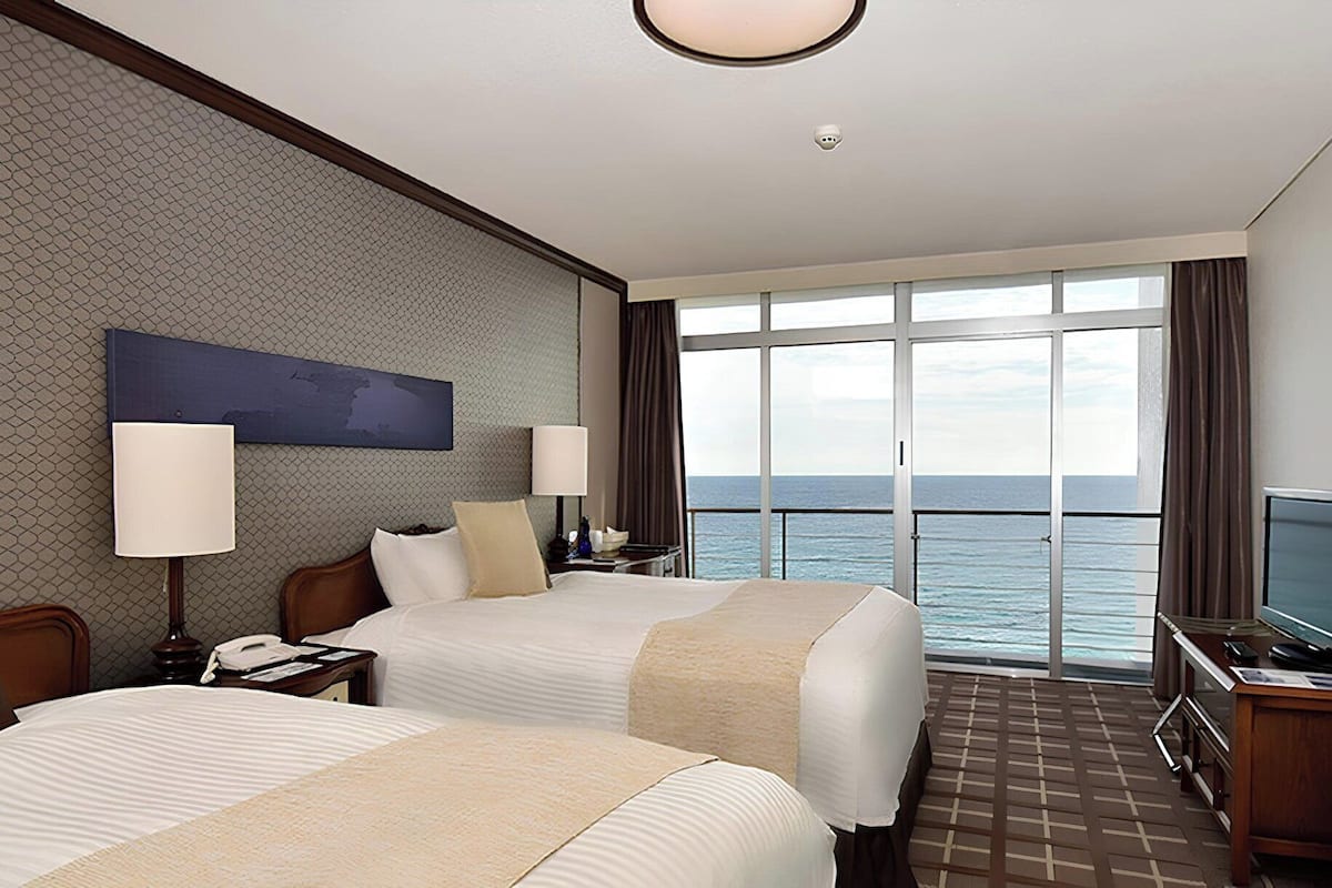 A hotel room with two beds, a large window with a view of the sea, a desk with a computer, and a TV.