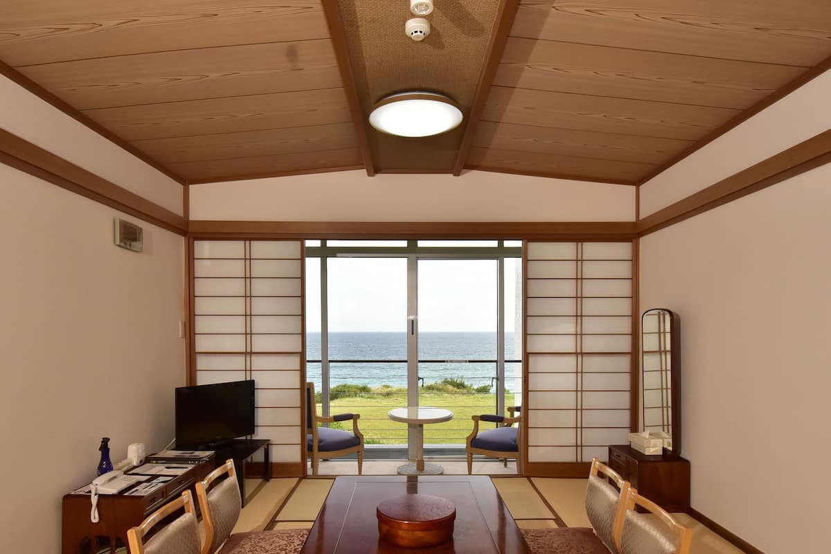 A traditional Japanese-style room with tatami flooring, a low table with cushions, a television, and a view of the ocean through large sliding doors.