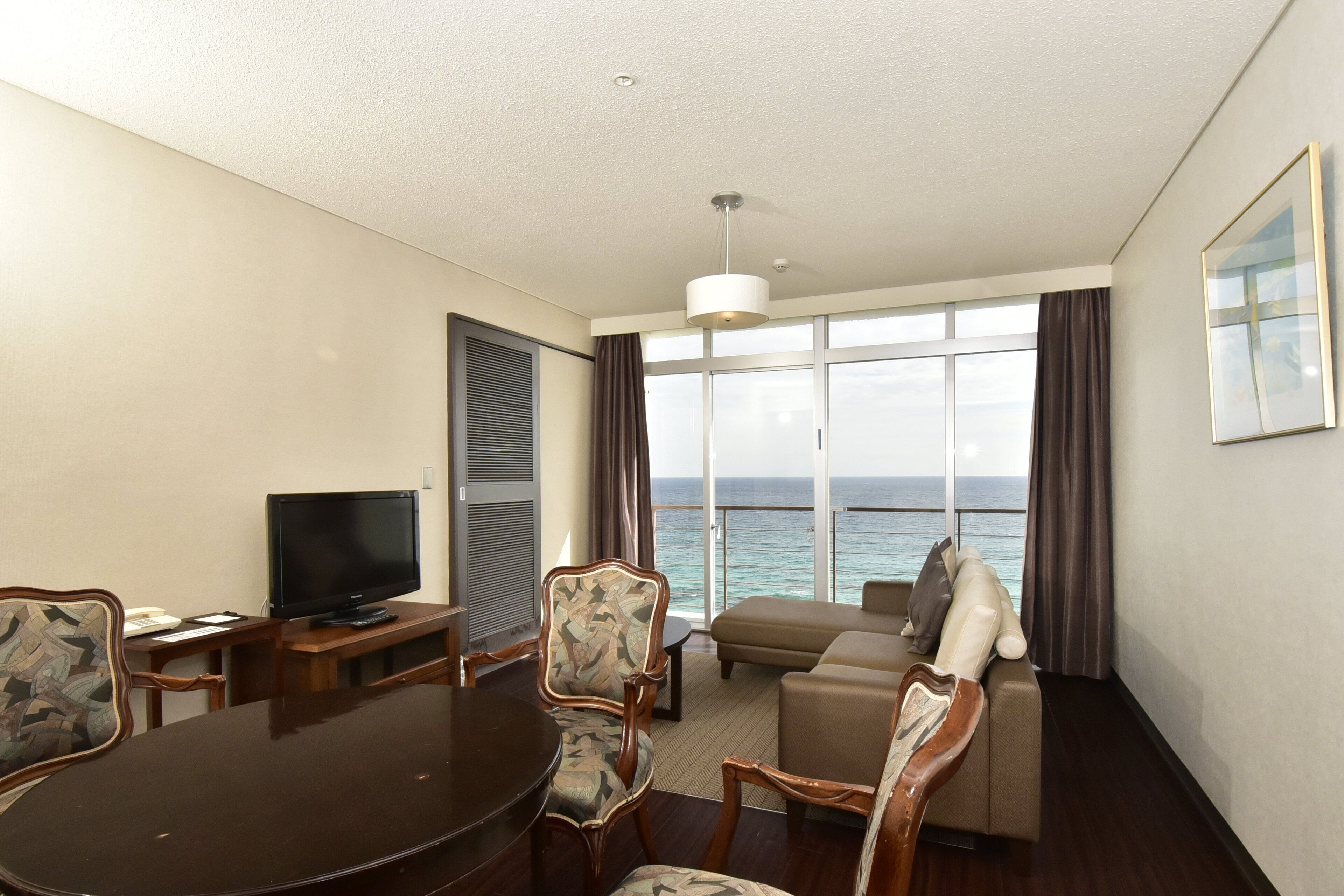 Habitación de hotel con una mesa de comedor, sillas, televisor, sofá y un ventanal con vista al mar.