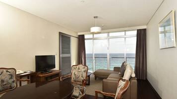Habitación de hotel con una mesa de comedor, sillas, televisor, sofá y un ventanal con vista al mar.