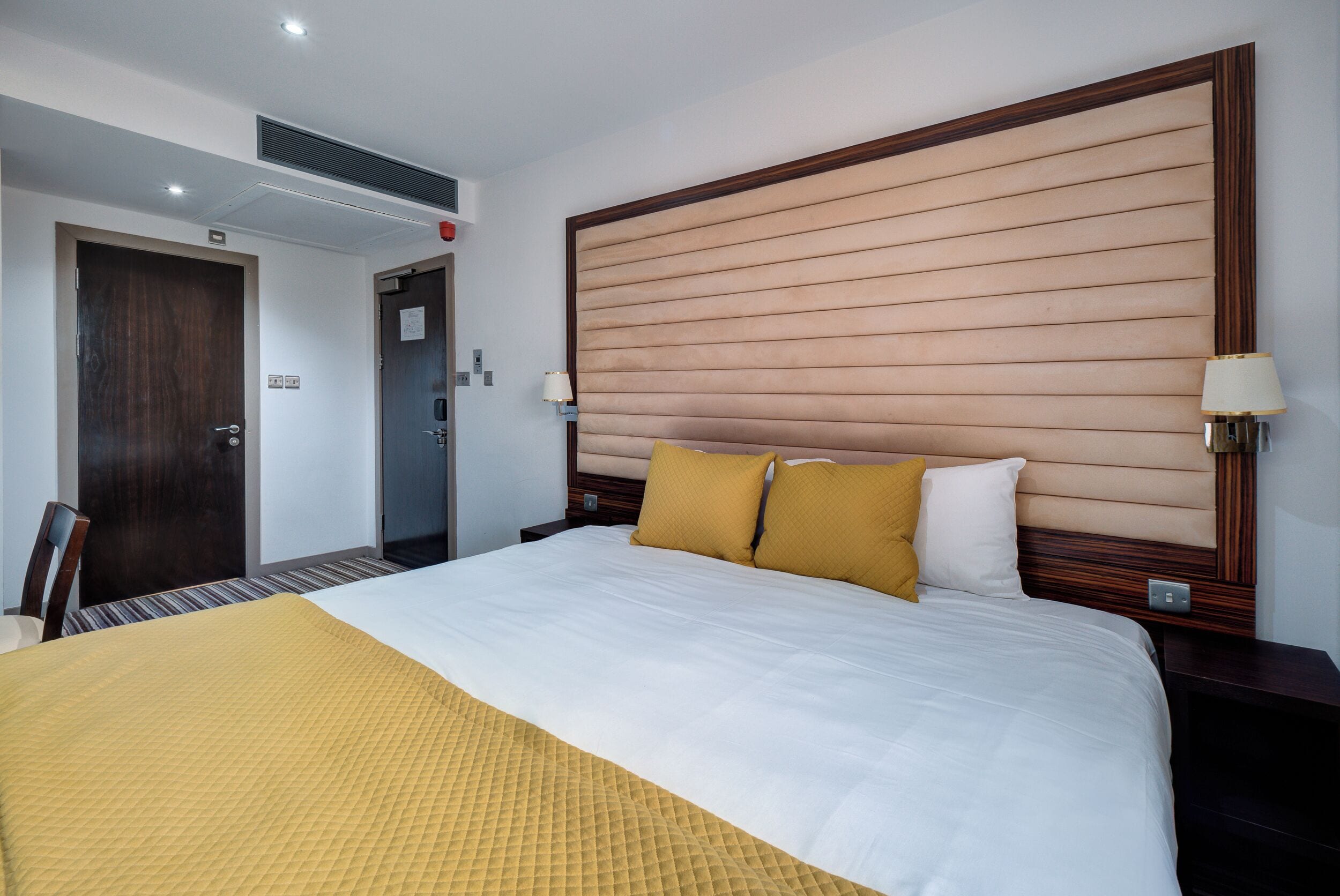 A hotel room with a large bed, wooden headboard, two bedside lamps, and a door with a sign.