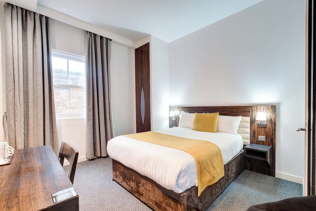 A hotel room with a large bed, a wooden headboard, a bedside table, a lamp, and a view of the city through the window.