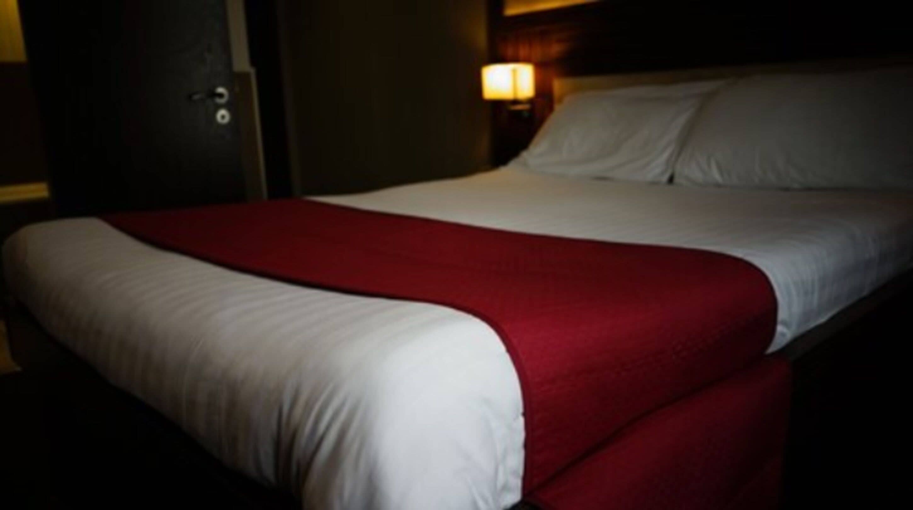 Un lit avec une couverture rouge et des draps blancs, une tête de lit en bois et une lampe fixée au mur.