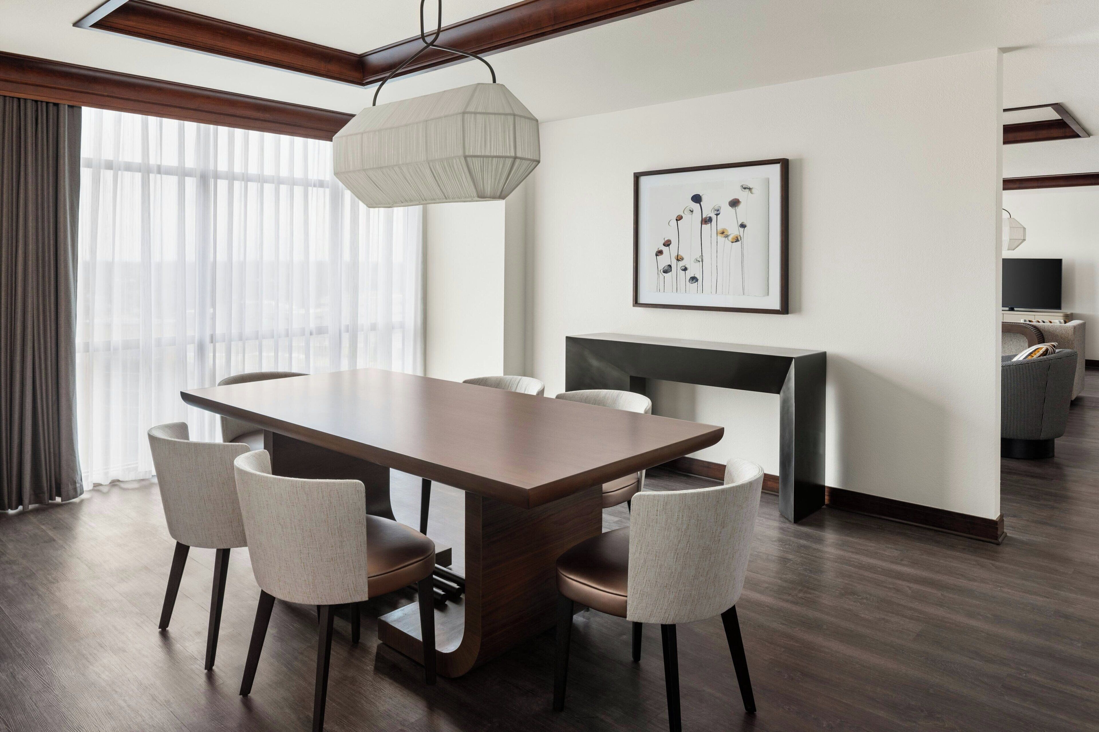 Un área de comedor moderna con una mesa de madera y seis sillas, un ventanal con cortinas translúcidas y una obra enmarcada en la pared.