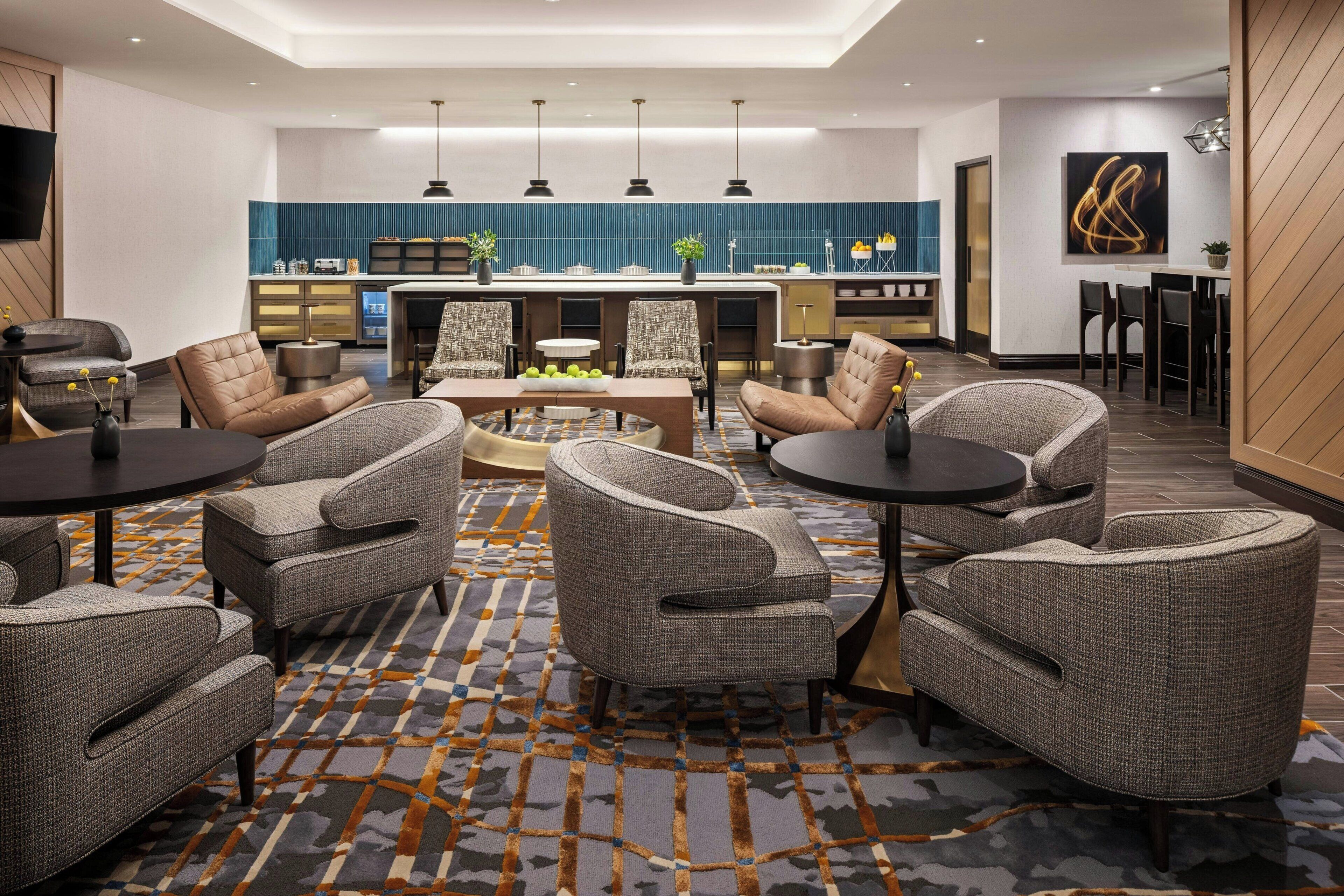 A modern hotel lobby with a central seating area, a kitchen in the background, and a dining area.