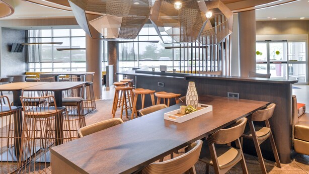 A modern restaurant with a long communal table, high stools, and a central bar area. - SpringHill Suites by Marriott Ashburn Dulles North (Ashburn)