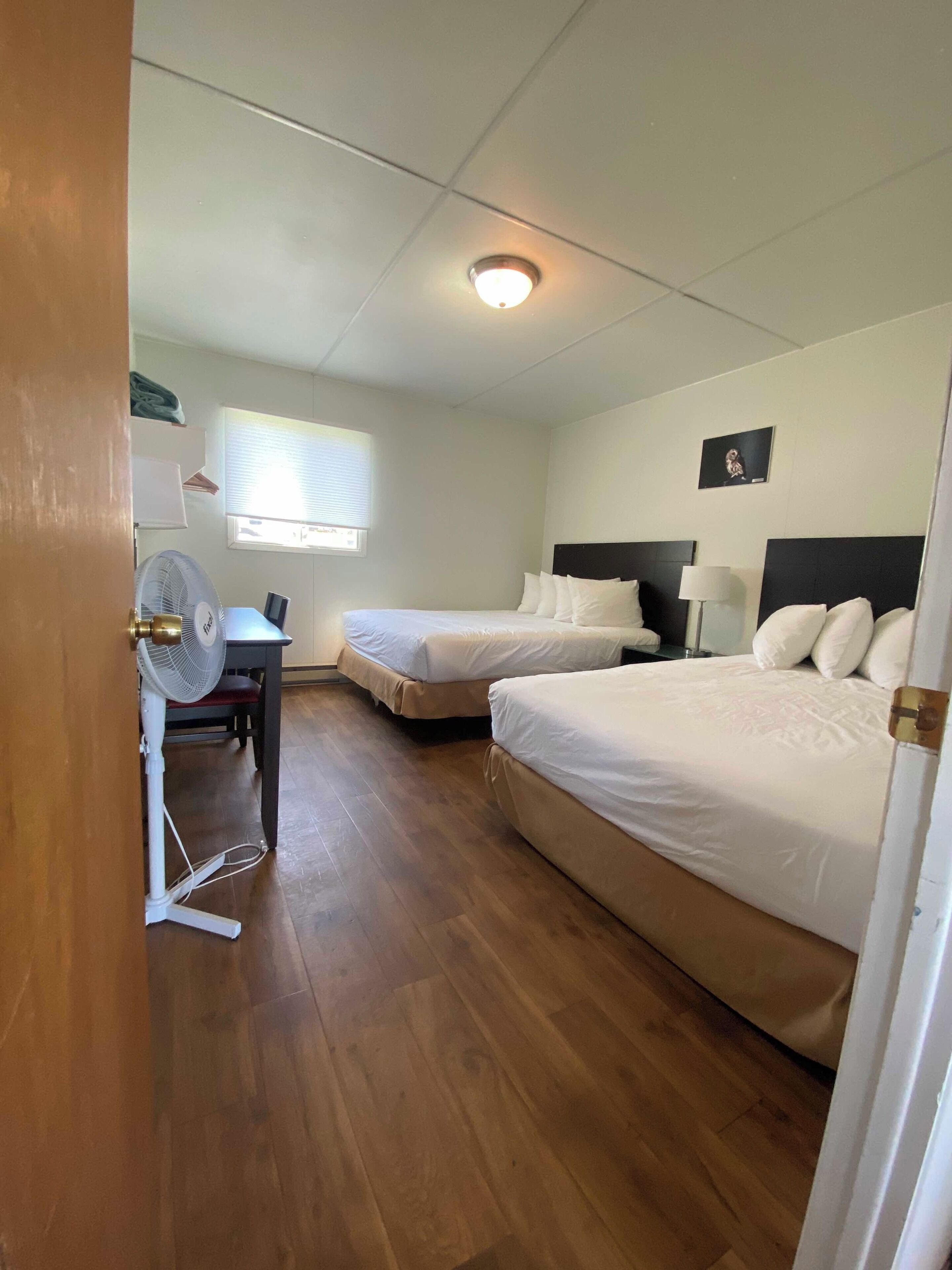 Habitación de hotel con dos camas, un escritorio, una silla, un ventilador de pie y una ventana con persianas.