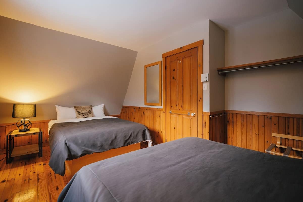 A room with two beds, a wooden door, a small table with a lamp, and a shelf above the door.