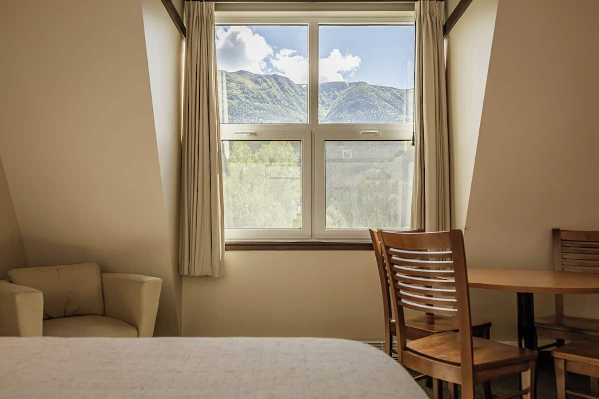 A bedroom with a bed, a wooden chair, a small table, and a window with a mountain view.