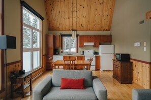 Een gezellig interieur van een chalet met een eethoek, keuken en een grijze bank met een rode kussen.