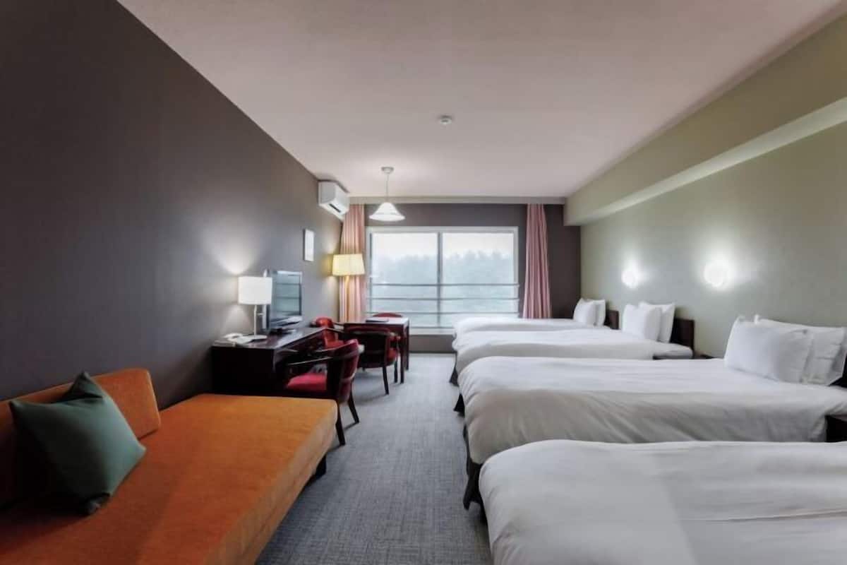A hotel room with three beds, a desk with a chair, a sofa, and a window with curtains.