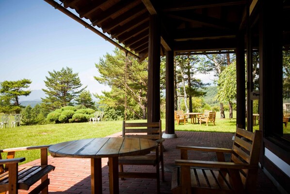 A covered outdoor seating area with a table and chairs, surrounded by a garden with trees and a view of the mountains. - Tsumagoi Prince Hotel (Tsumagoi)