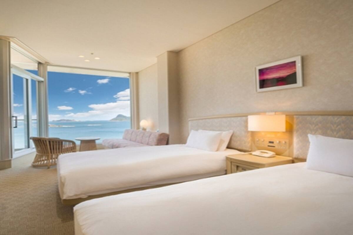 Une chambre d’hôtel avec deux lits, une vue sur la mer, une chaise et une table de chevet avec une lampe.