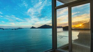 Une pièce dotée d’une grande fenêtre donnant sur une mer calme et des montagnes lointaines, le tout baigné par les couleurs du coucher de soleil.
