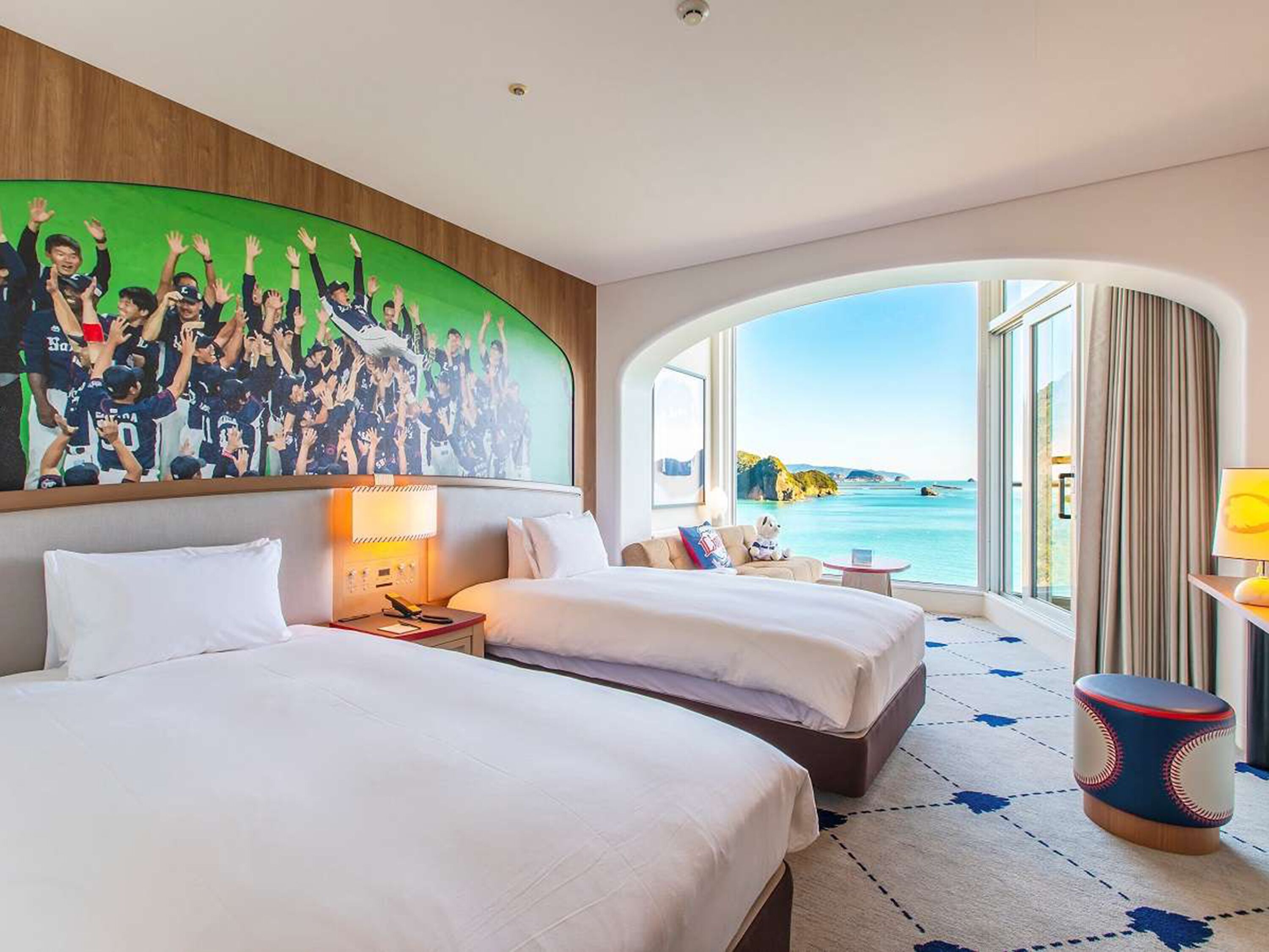 A hotel room with two beds, a large mural, a view of the sea, and a round table with a baseball-themed ottoman.