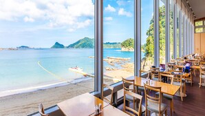 Un restaurant doté de grandes fenêtres donnant sur une plage et la mer, de tables et de chaises en bois, et offrant une vue sur des îles lointaines.
