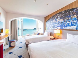 Habitación de hotel con dos camas, un ventanal con vista al mar, un televisor y un cuadro de un equipo deportivo.