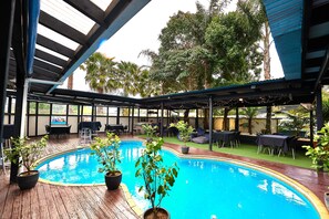 An indoor swimming pool with a wooden deck, surrounded by outdoor seating and potted plants. - Mount Richmond Hotel (Auckland)