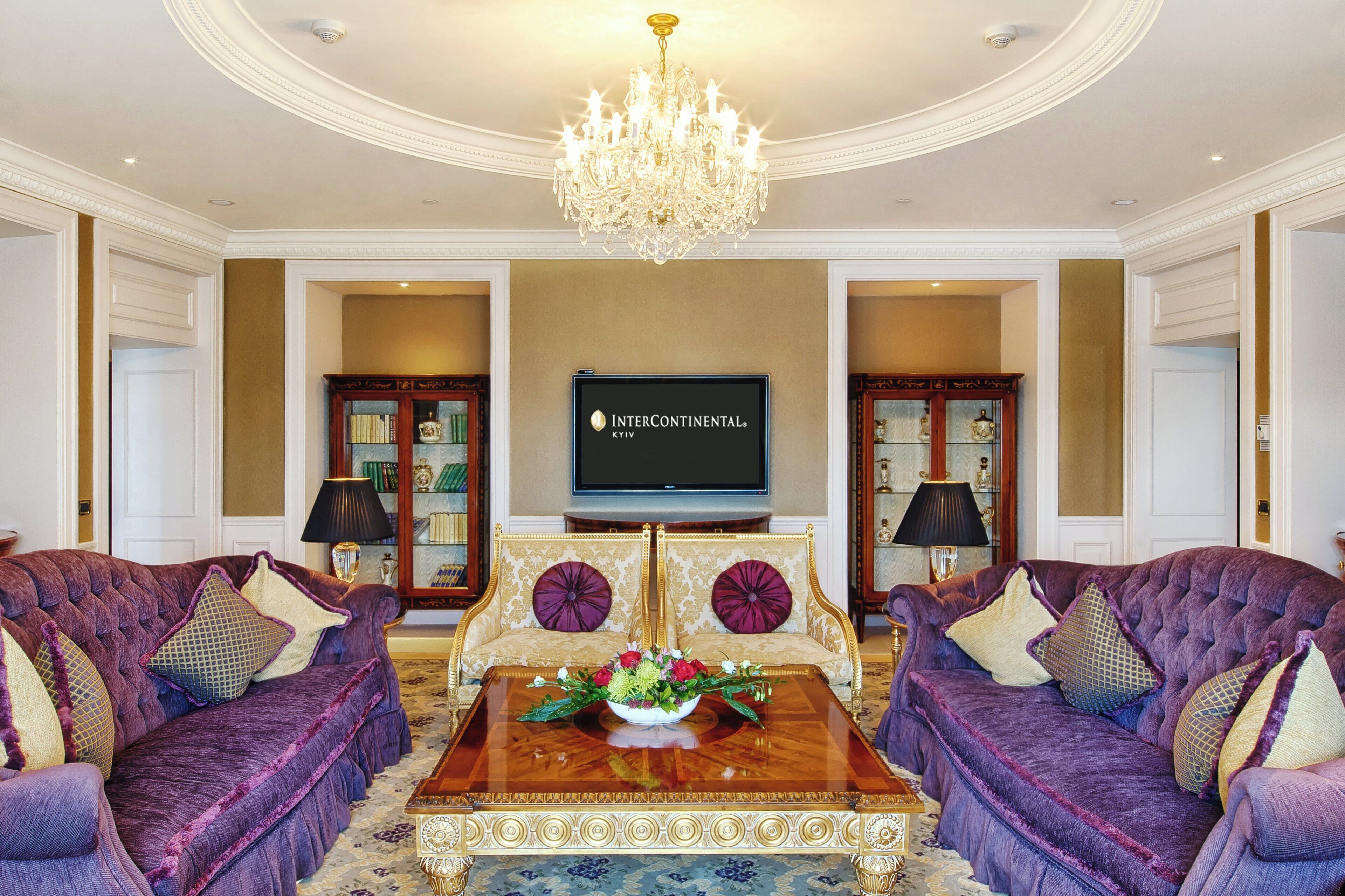 A luxurious living room with a chandelier, two sofas, a coffee table with a floral arrangement, and a flat-screen TV mounted on the wall.