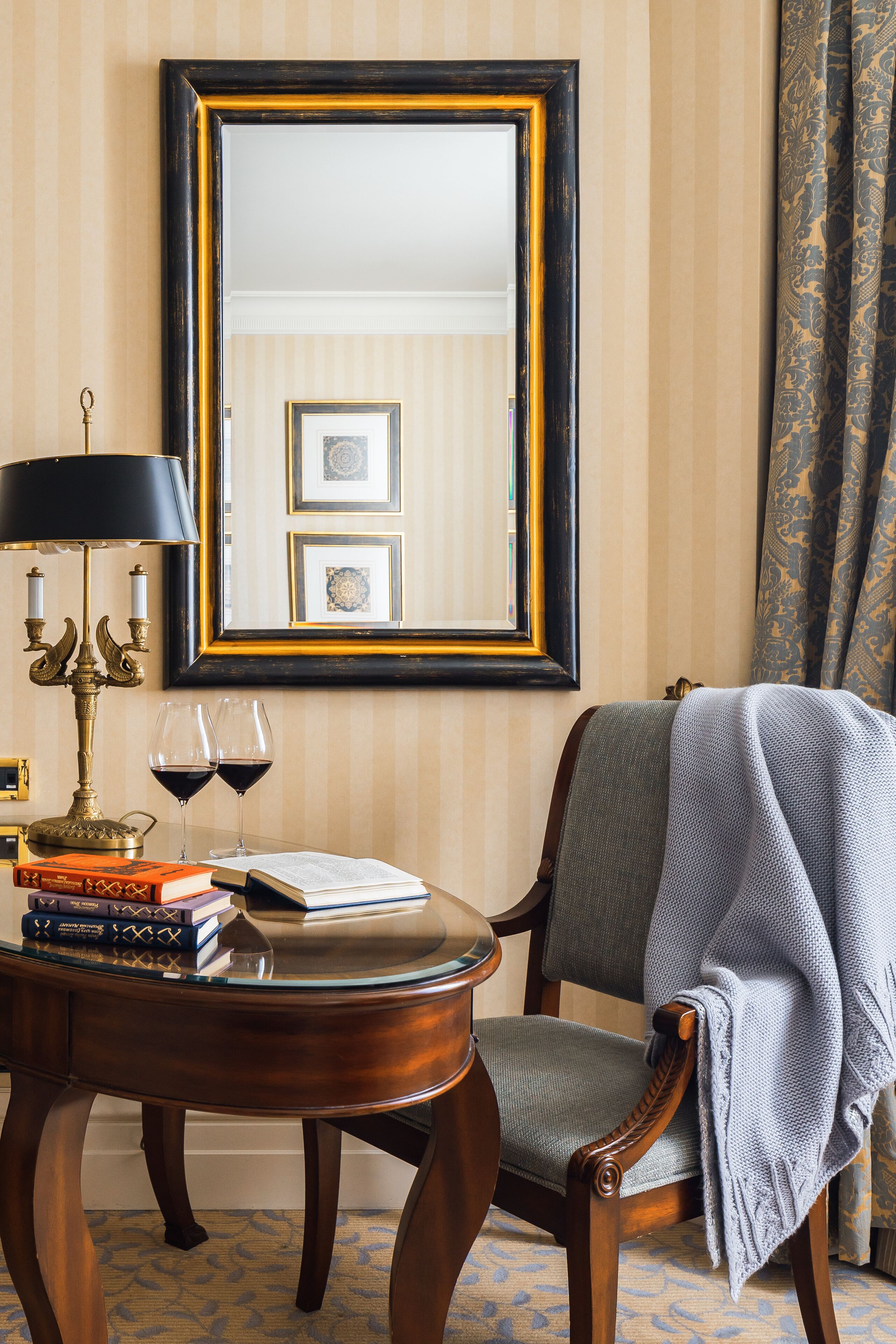 Une pièce avec une chaise en bois, une table sur laquelle se trouvent des livres et des lunettes, une lampe et un miroir accroché au mur.