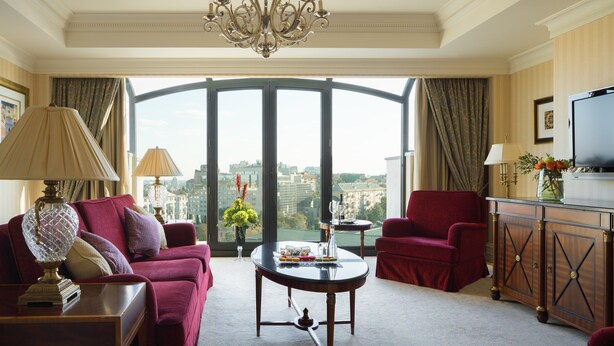 A luxurious room with a large window, a chandelier, a red sofa, a red armchair, a coffee table with snacks, and a TV on a cabinet. - InterContinental Kyiv by IHG (Kyiv)