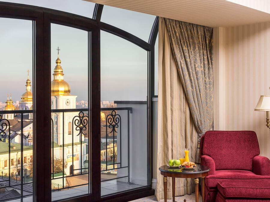 Habitación con un ventanal que ofrece vistas a cúpulas doradas y un paisaje urbano. Hay un sillón rojo y una mesita con fruta y una lámpara.