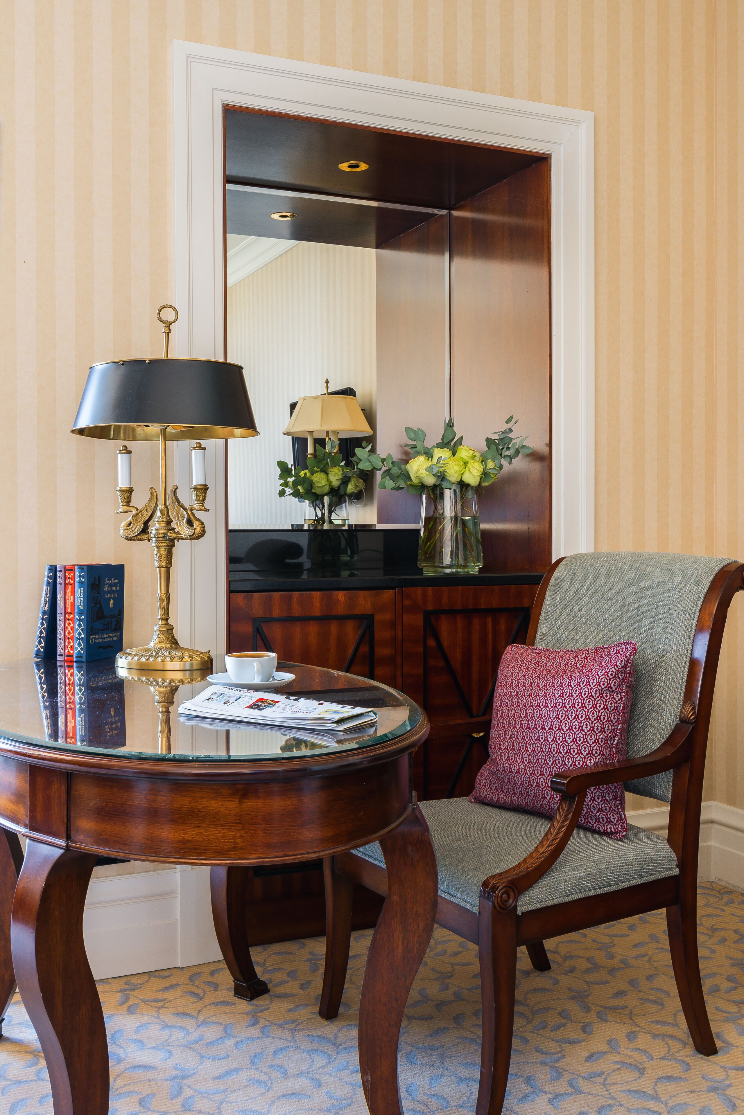 A wooden chair with a cushion, a round table with a lamp, books, and a vase with flowers.