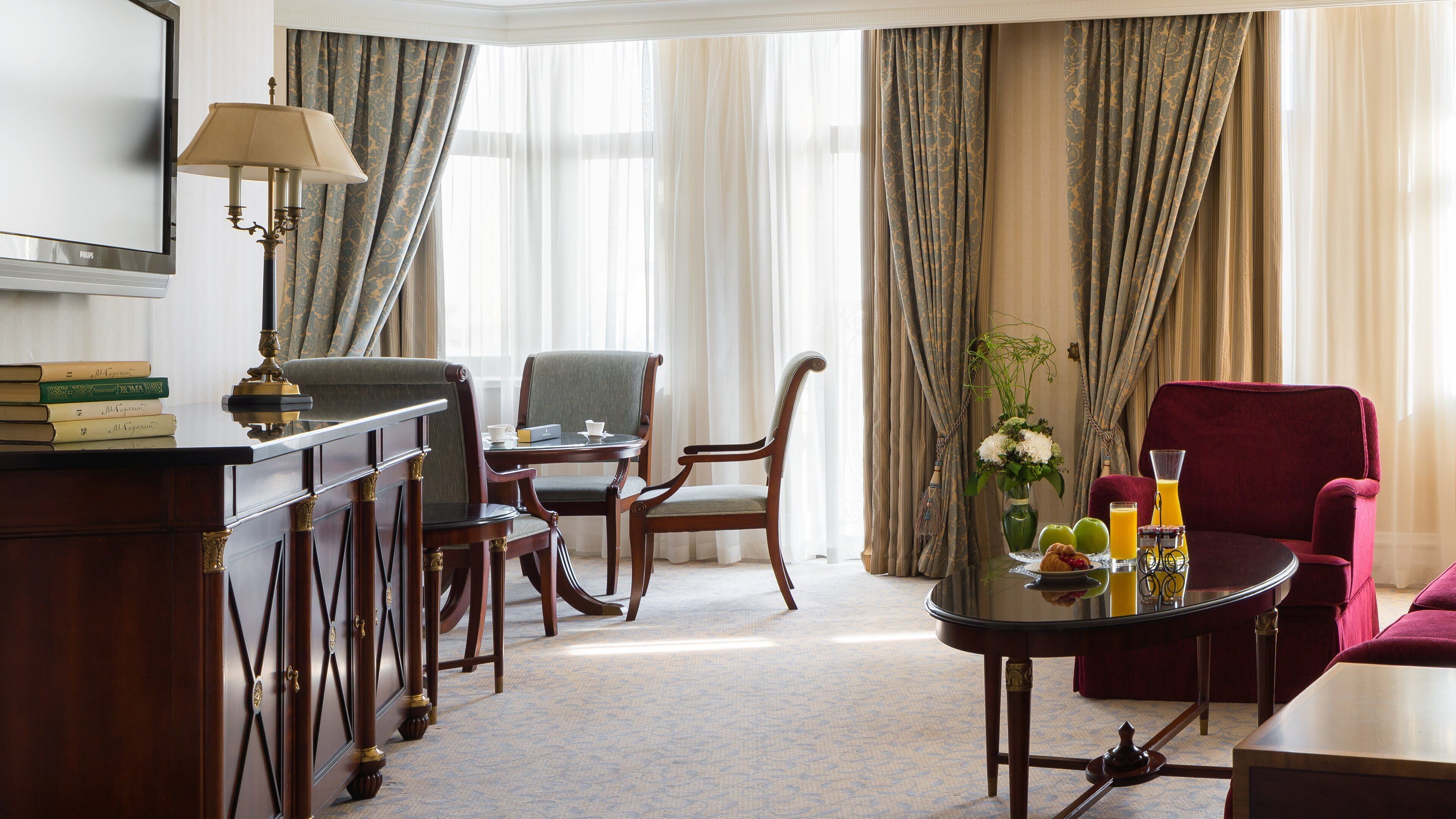 Habitación de hotel con escritorio, dos sillas, un sillón rojo, una mesa de comedor y elementos para el desayuno, además de una lámpara.