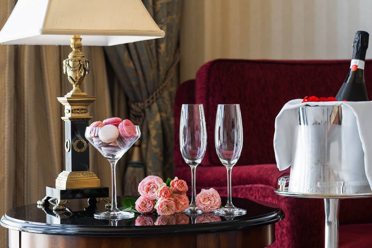 A table with a lamp, two glasses, a bottle of champagne, a macaron-filled glass, and pink roses.