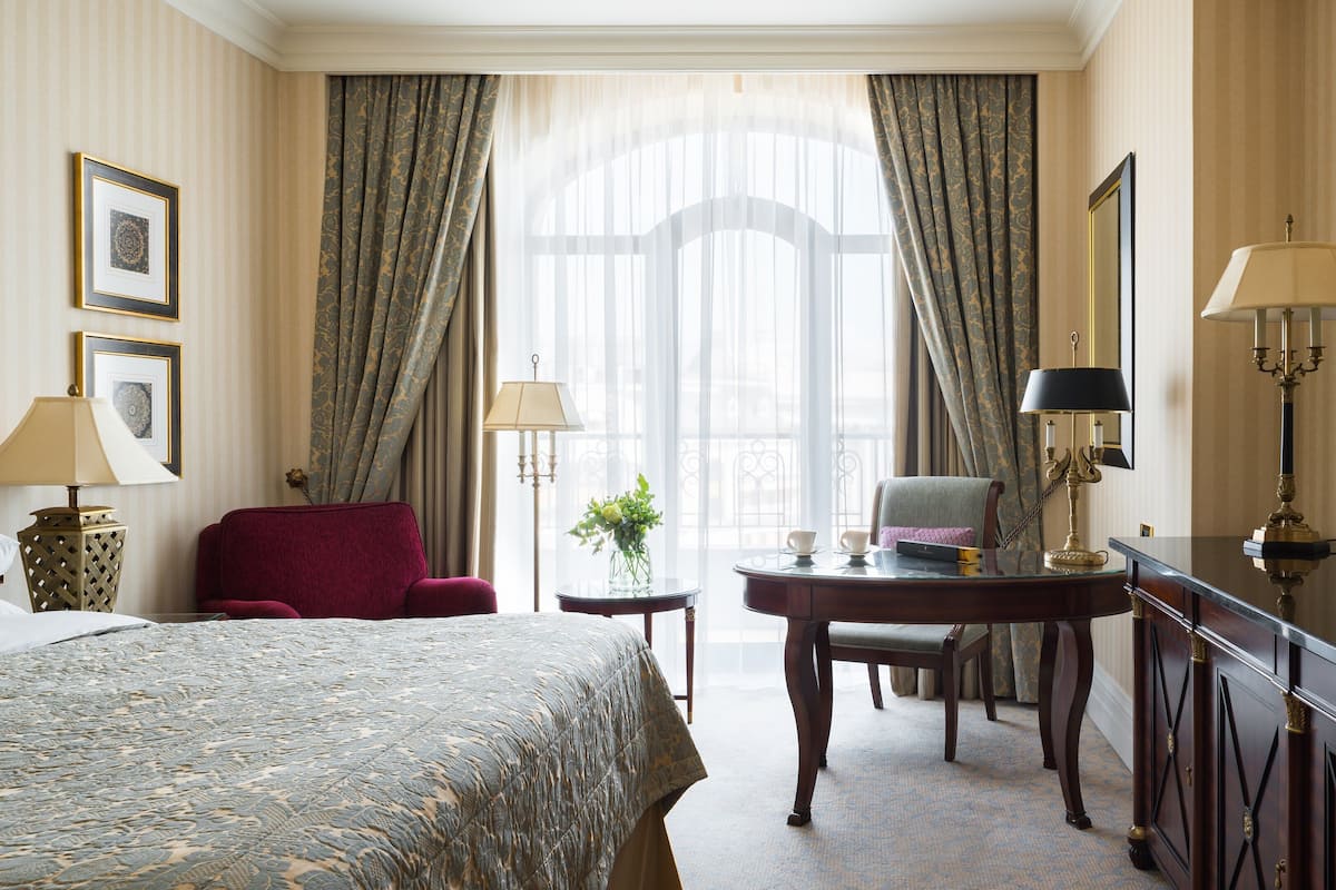 A hotel room with a large bed, a desk with a chair, a small table with a vase of flowers, and a large window with curtains.
