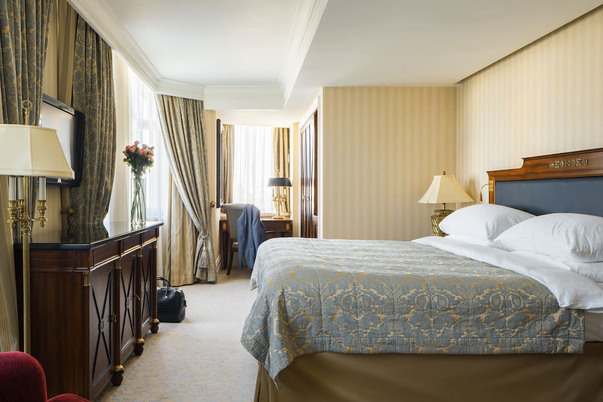 A hotel room with a large bed, two bedside lamps, a wooden nightstand, a desk with a lamp, a chair, and a wardrobe.