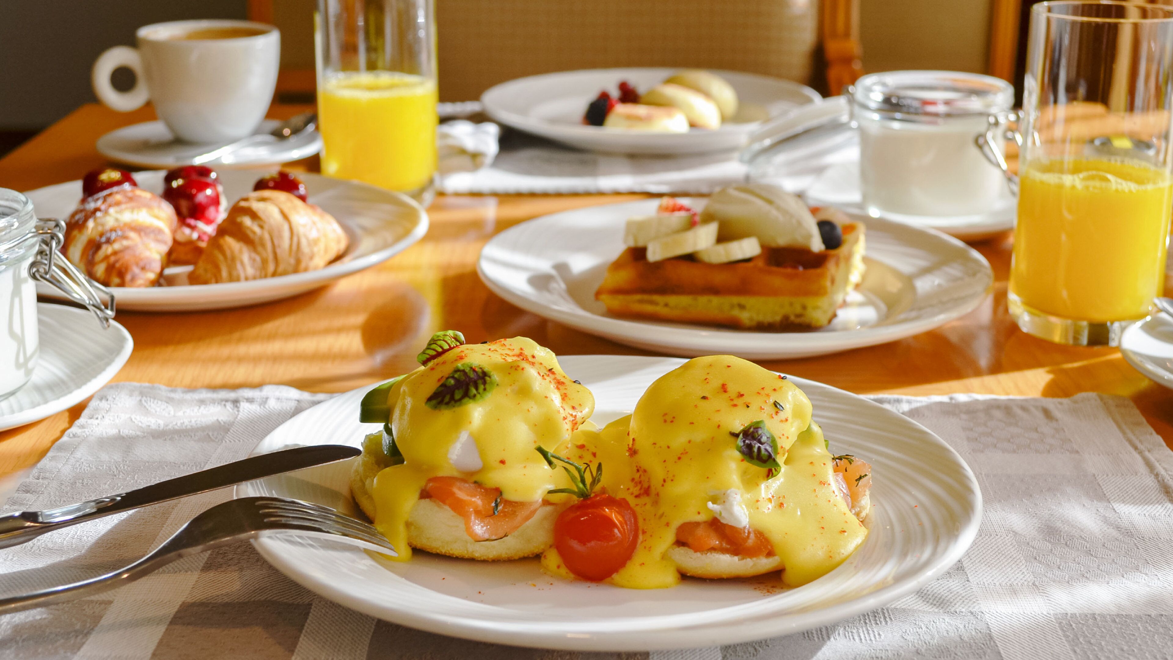 A breakfast setting with plates of food, including eggs Benedict and pastries, along with glasses of orange juice and a cup of coffee.