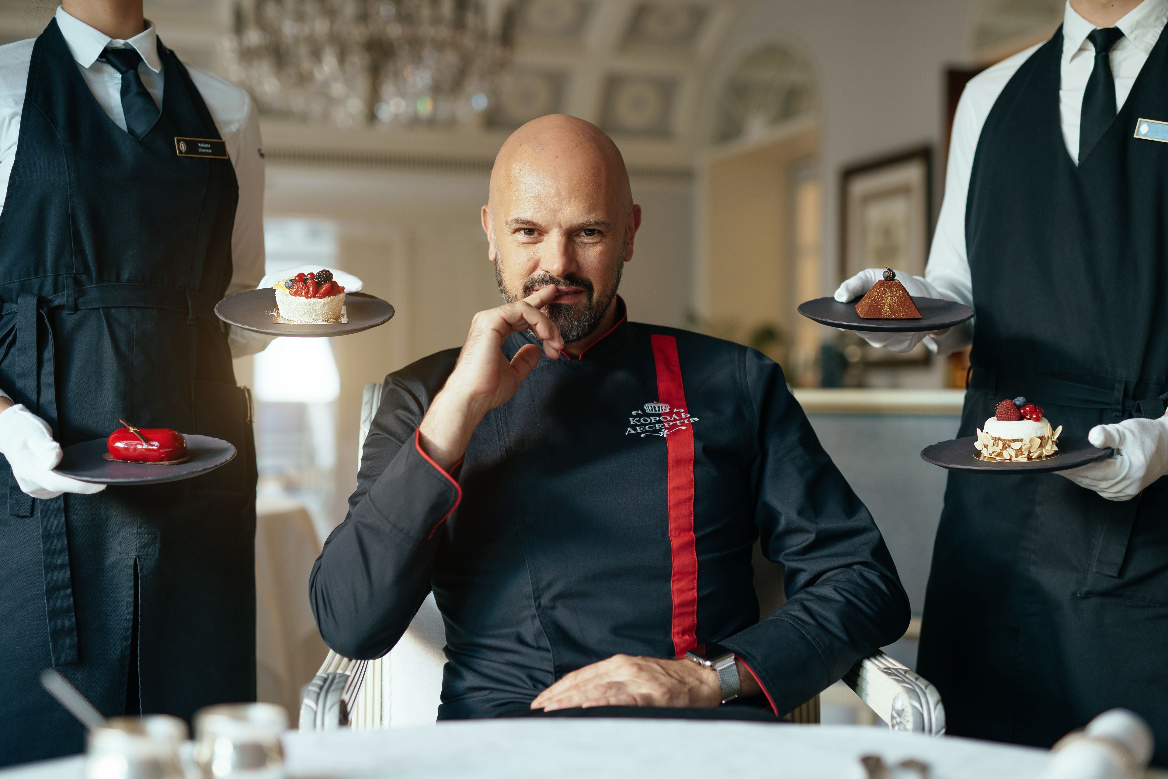 A person in a black chef's jacket with a red stripe, seated at a table with a dessert in front of them.