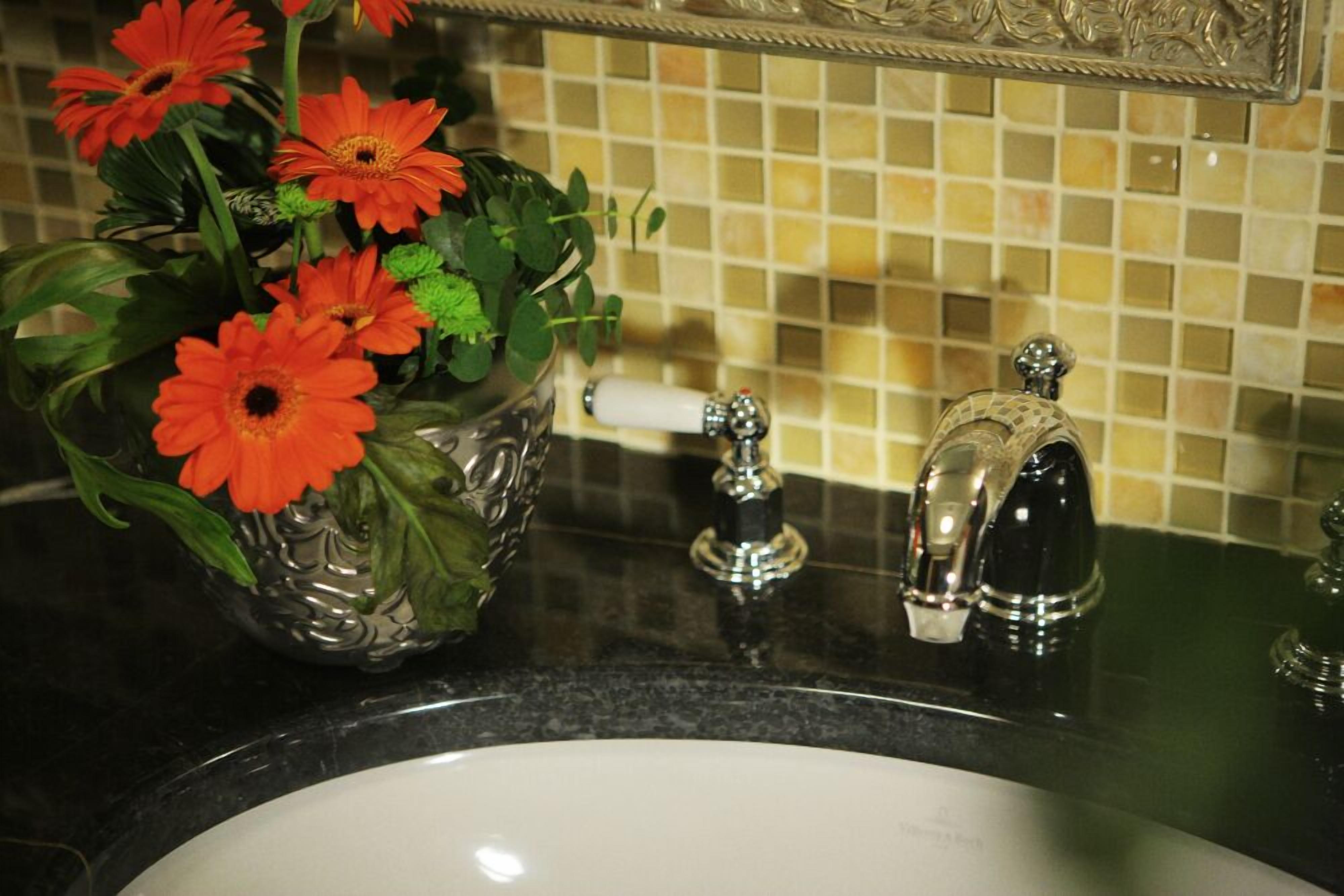 A bathroom with a sink, a decorative glass vase with flowers, a mirror, and a tiled backsplash.