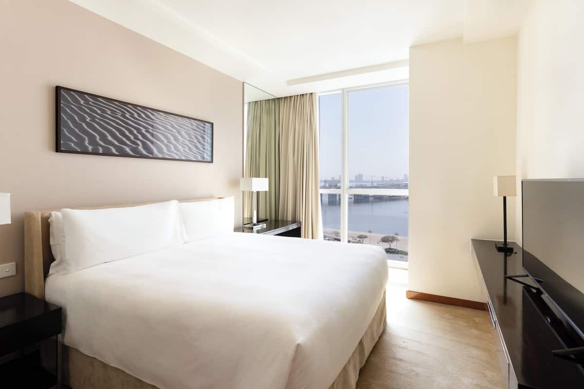Une chambre d’hôtel avec un grand lit, un téléviseur à écran plat, une table de chevet, une lampe, une fenêtre donnant sur un plan d’eau et un tableau au mur.