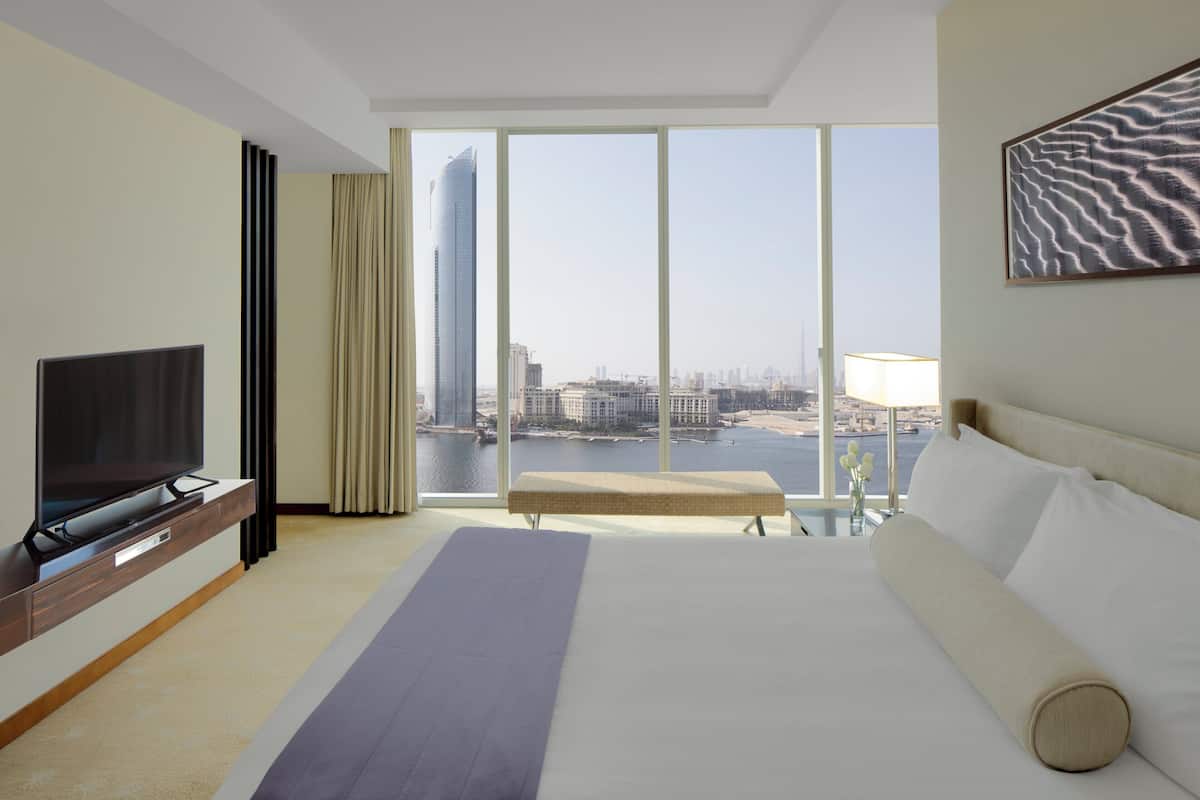 Une chambre d’hôtel dotée d’un grand lit, d’un téléviseur à écran plat, d’un banc et offrant une vue sur le paysage urbain grâce à de grandes fenêtres.