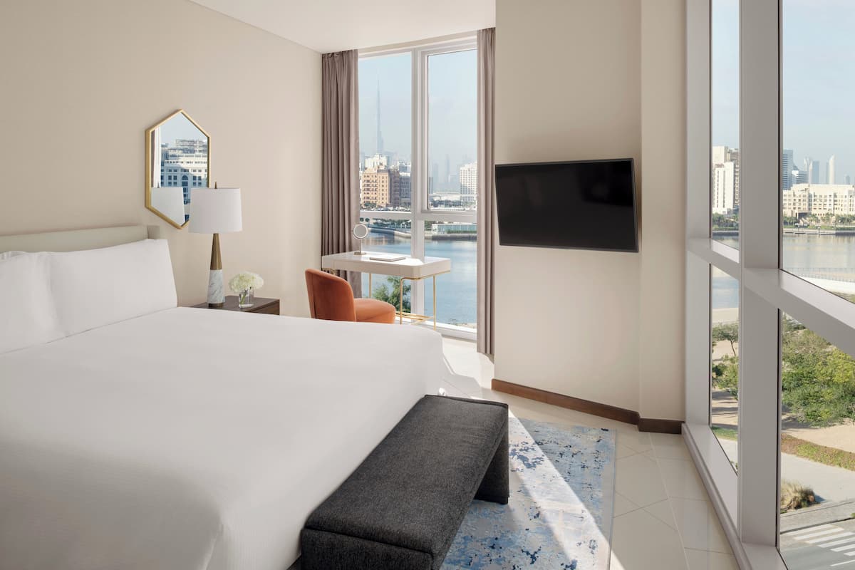 Une chambre d’hôtel dotée d’un grand lit, d’un bureau avec une chaise, d’un téléviseur fixé au mur et offrant une vue sur le paysage urbain à travers de grandes fenêtres.