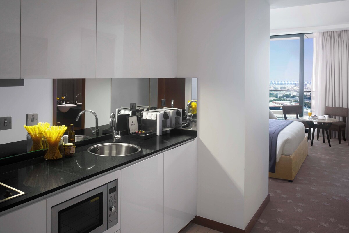Eine moderne Hotelküche mit Spüle, einer Kaffeemaschine und einem Toaster. Im Hintergrund sind ein Bett und ein Essbereich zu sehen.