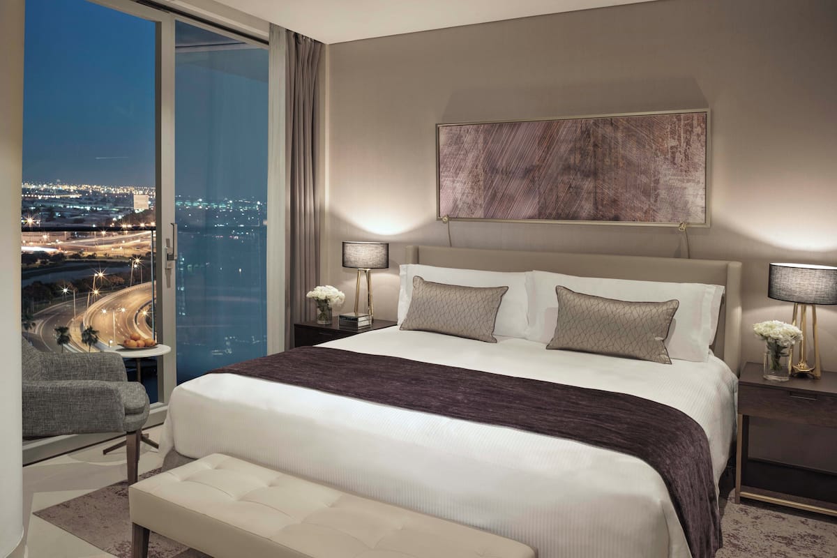 Une chambre d’hôtel moderne avec un grand lit, une banquette, une table de chevet avec une lampe et une vue sur le paysage urbain par une grande fenêtre.
