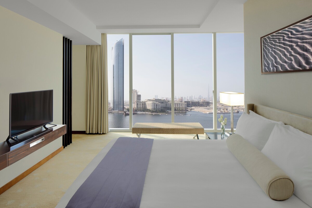 Ein Hotelzimmer mit einem großen Bett, einem Flachbildfernseher, einer Bank und Blick auf eine Stadtlandschaft durch große Fenster.