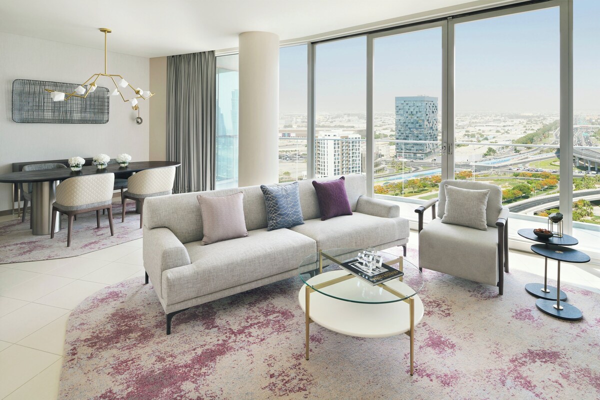 Ein modernes Wohnzimmer mit Sofa, Sessel und Couchtisch, großen Fenstern mit Stadtblick und einem Essbereich mit Stühlen und Tisch.