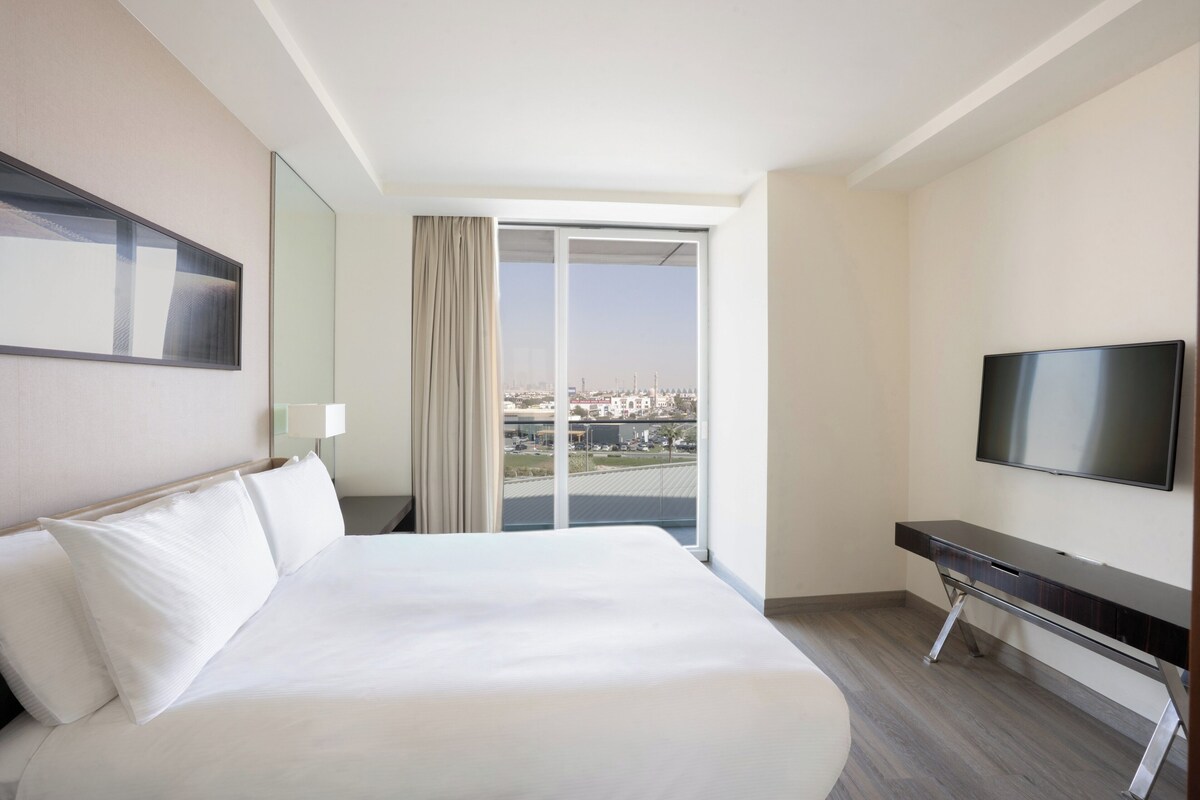 Ein Hotelzimmer mit einem großen Bett, einem Fernseher, einem Schreibtisch und Blick auf die Stadt durch das Fenster.