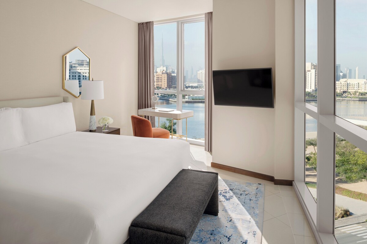Ein Hotelzimmer mit einem großen Bett, einem Schreibtisch mit Stuhl, einem an der Wand befestigten Fernseher und Blick auf eine Stadtlandschaft durch große Fenster.