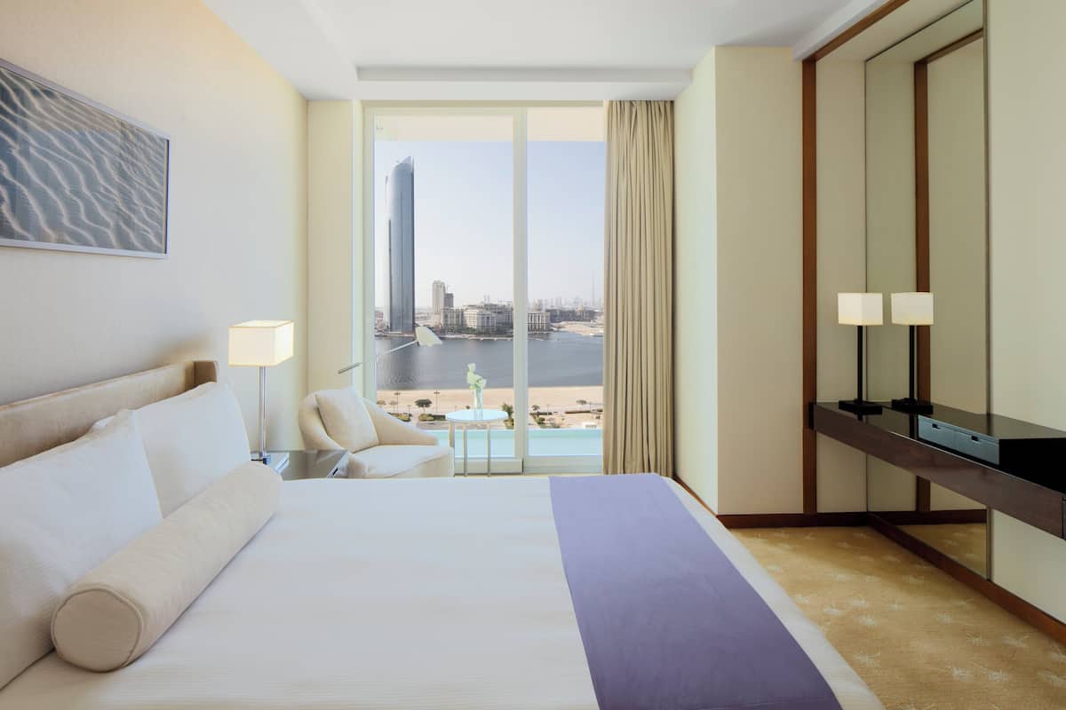 Une chambre d’hôtel avec un grand lit, un bureau avec une lampe, une chaise et une vue sur le paysage urbain et l’eau.