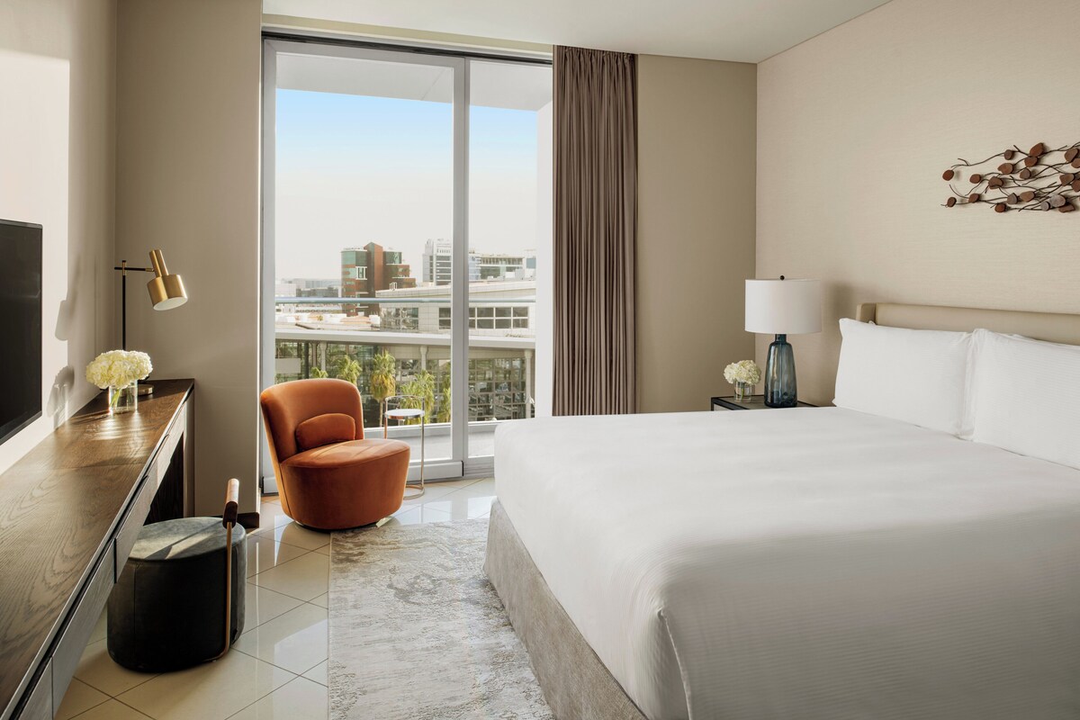 Ein Hotelzimmer mit einem großen Bett, einem Schreibtisch mit Fernseher, einem Sessel und Blick auf die Stadt durch große Fenster.
