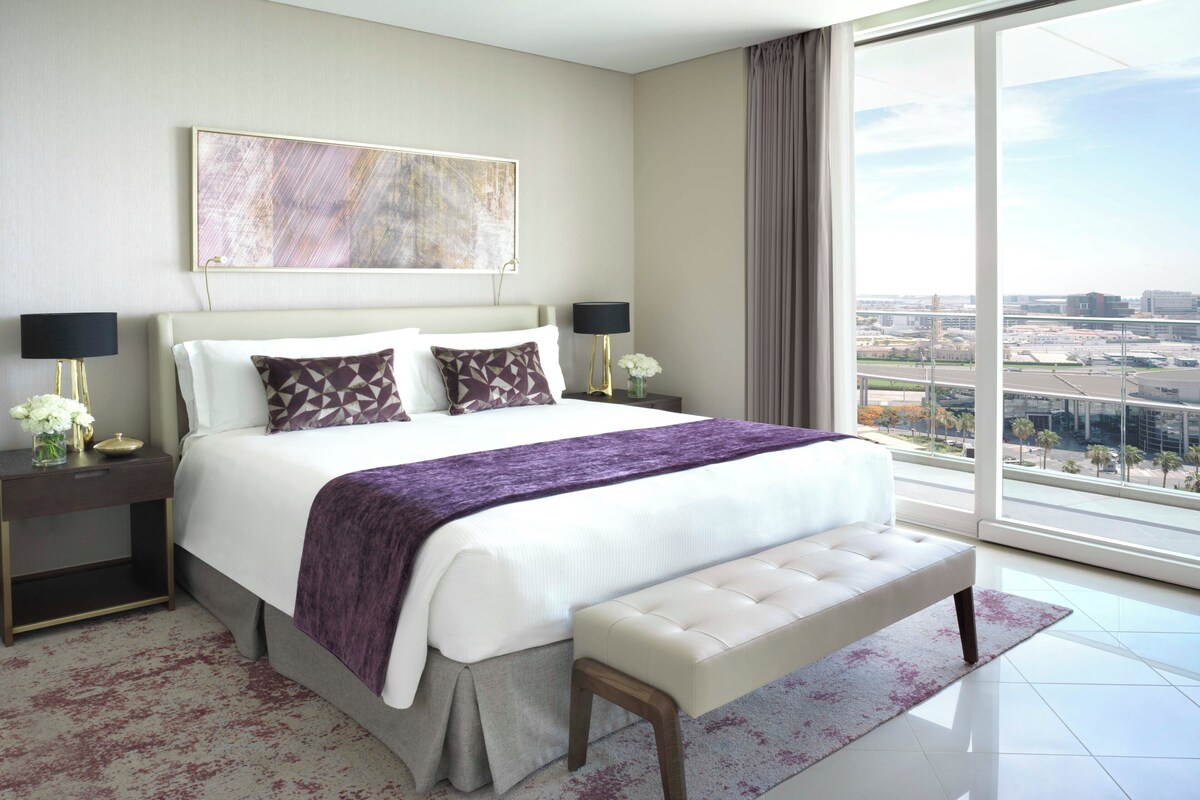 A modern hotel room with a large bed, two bedside tables with lamps, a bench, and a large window offering a city view.