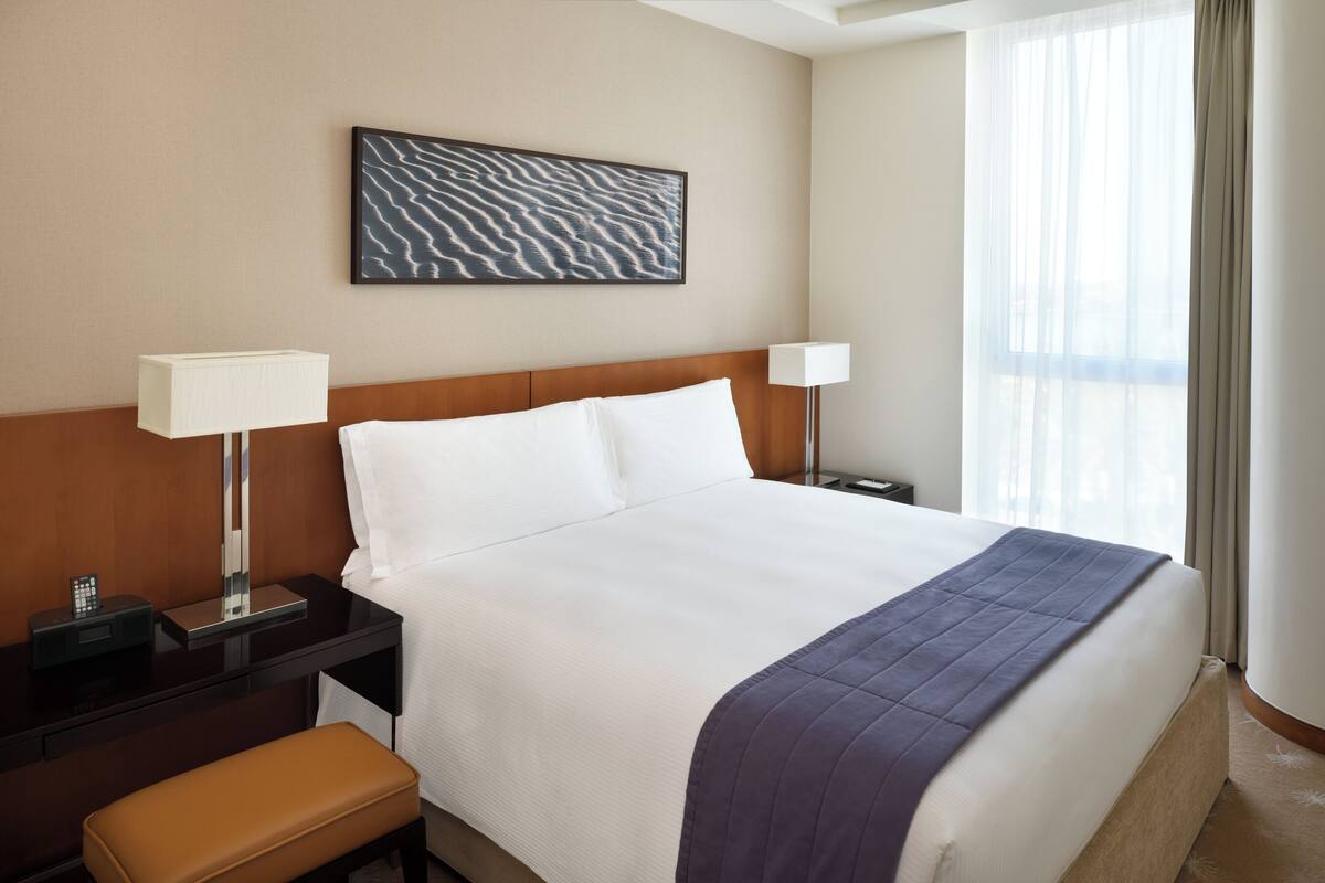 Une chambre d’hôtel avec un grand lit, deux tables de chevet, une petite table, un banc, une œuvre d’art accrochée au mur et une fenêtre avec des rideaux.