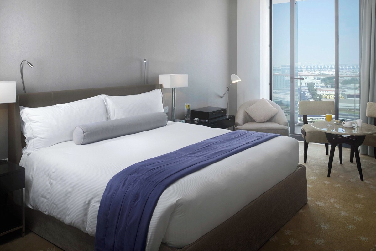Ein Hotelzimmer mit einem großen Bett, einem Schreibtisch mit Stuhl, einem Fernseher und Blick auf die Stadt.
