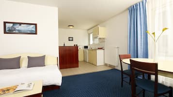 A hotel room with a bed, a desk, a chair, a small kitchen area, and a window with curtains.